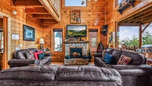 A cozy cabin interior with a stone fireplace, leather sofas, wooden walls, and a ceiling fan.