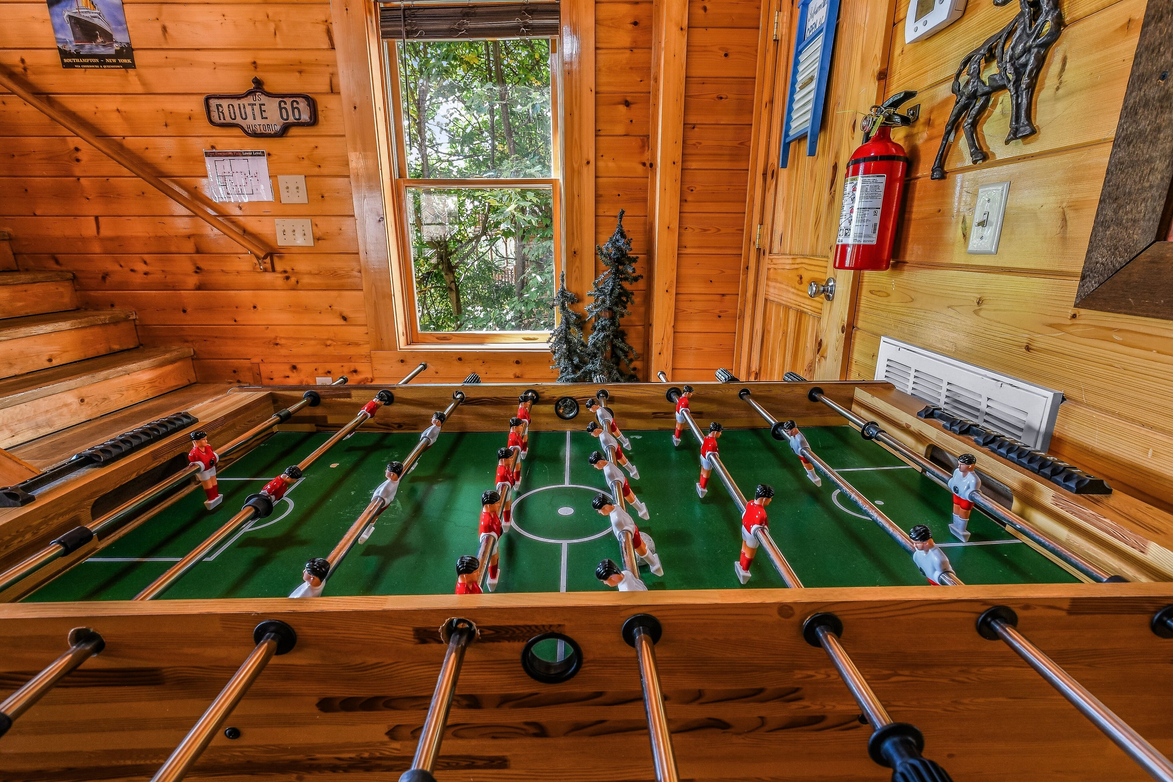 Un interior de madera con una mesa de fútbol de mesa, una ventana con vistas a árboles y un extintor en la pared.