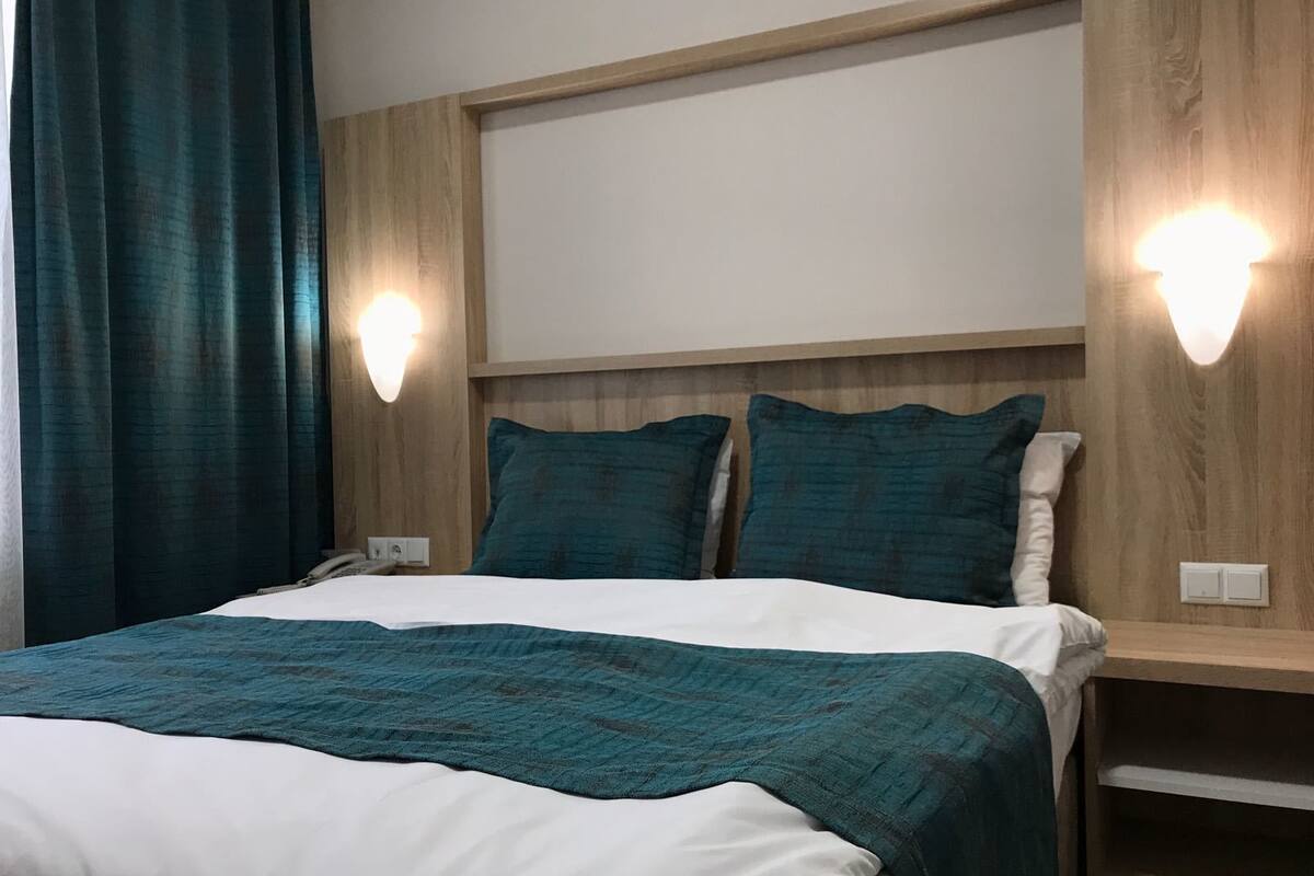 Habitación de hotel con una cama, dos luces fijadas a la pared, cabecero de madera y una manta color turquesa.