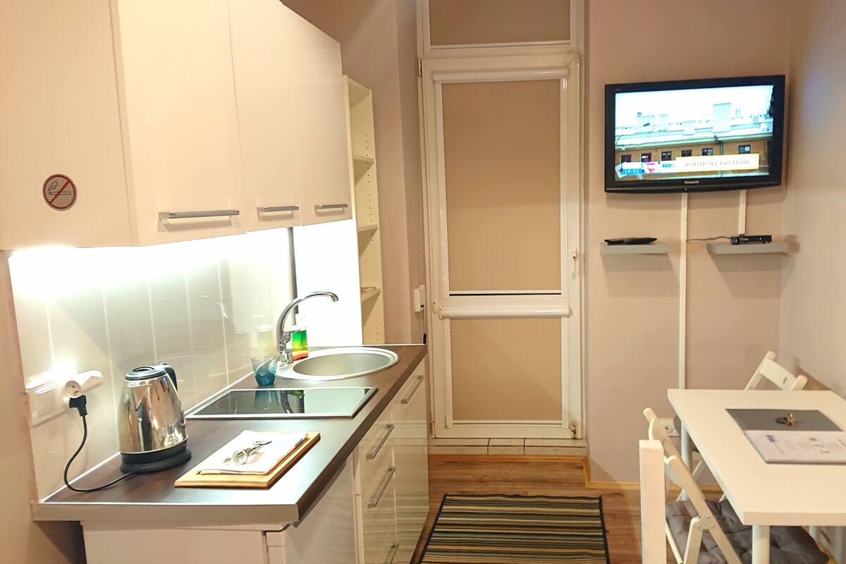 A compact kitchenette with a sink, a kettle, and a small table with a chair. A television is mounted on the wall, and a door leads to another room.