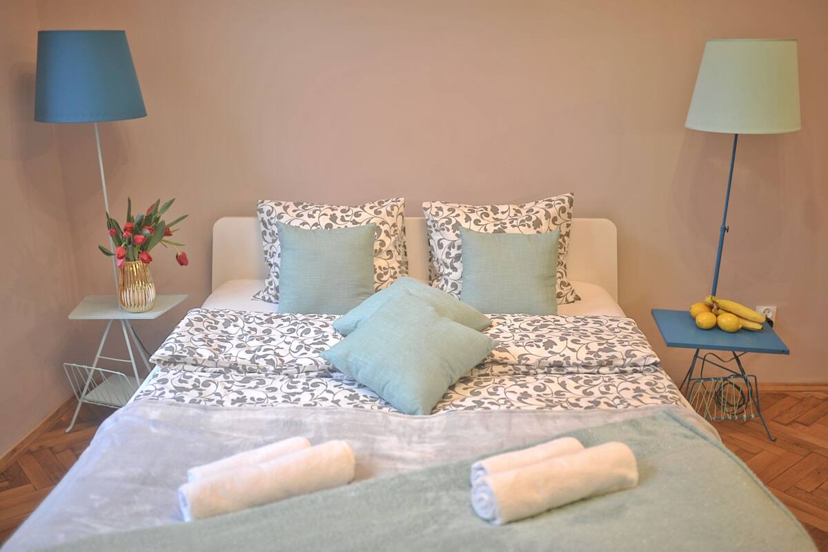 A neatly made bed with patterned bedding, two bedside lamps, a small table with a vase of flowers, and a banana on a side table.