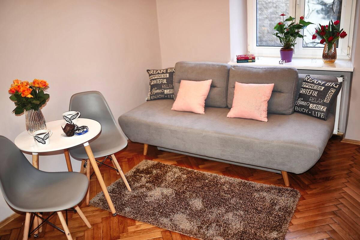 A living room with a grey sofa, two chairs, a small round table, and a brown rug. There are two potted plants on the windowsill.