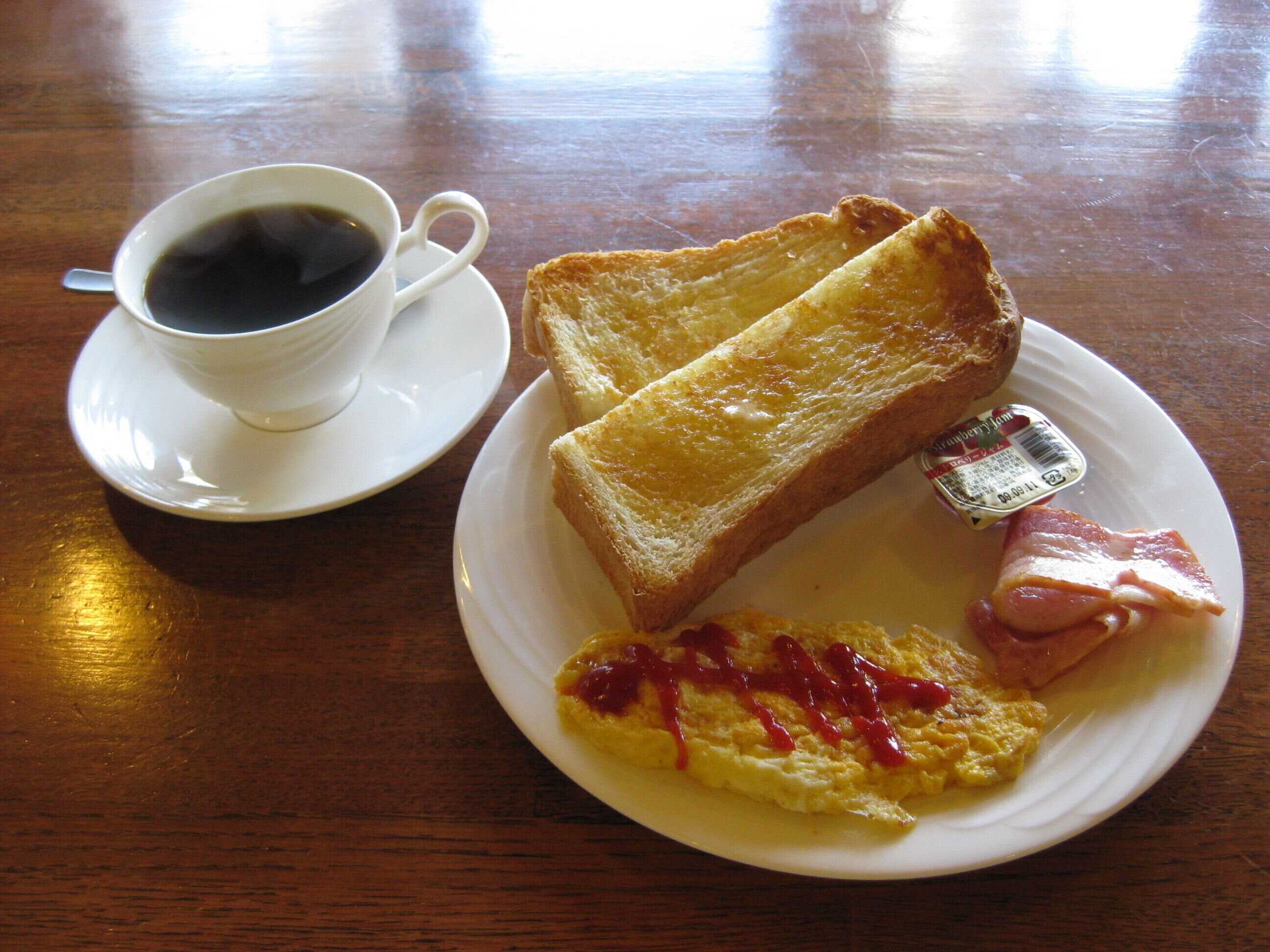 A breakfast plate with toast, ham, scrambled eggs, and ketchup, and a cup of coffee on a saucer.