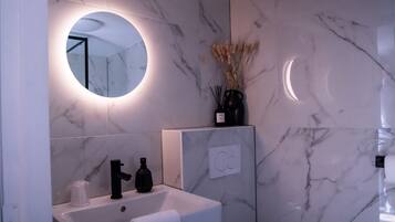 A bathroom with a white sink, black faucet, and a round mirror with a light. There is a vase with dried plants on the wall.