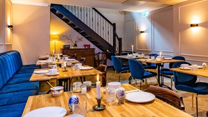 A dining area with wooden tables, blue upholstered seating, and a staircase in the background. - Hotel Janssen (Valkenburg aan de Geul)