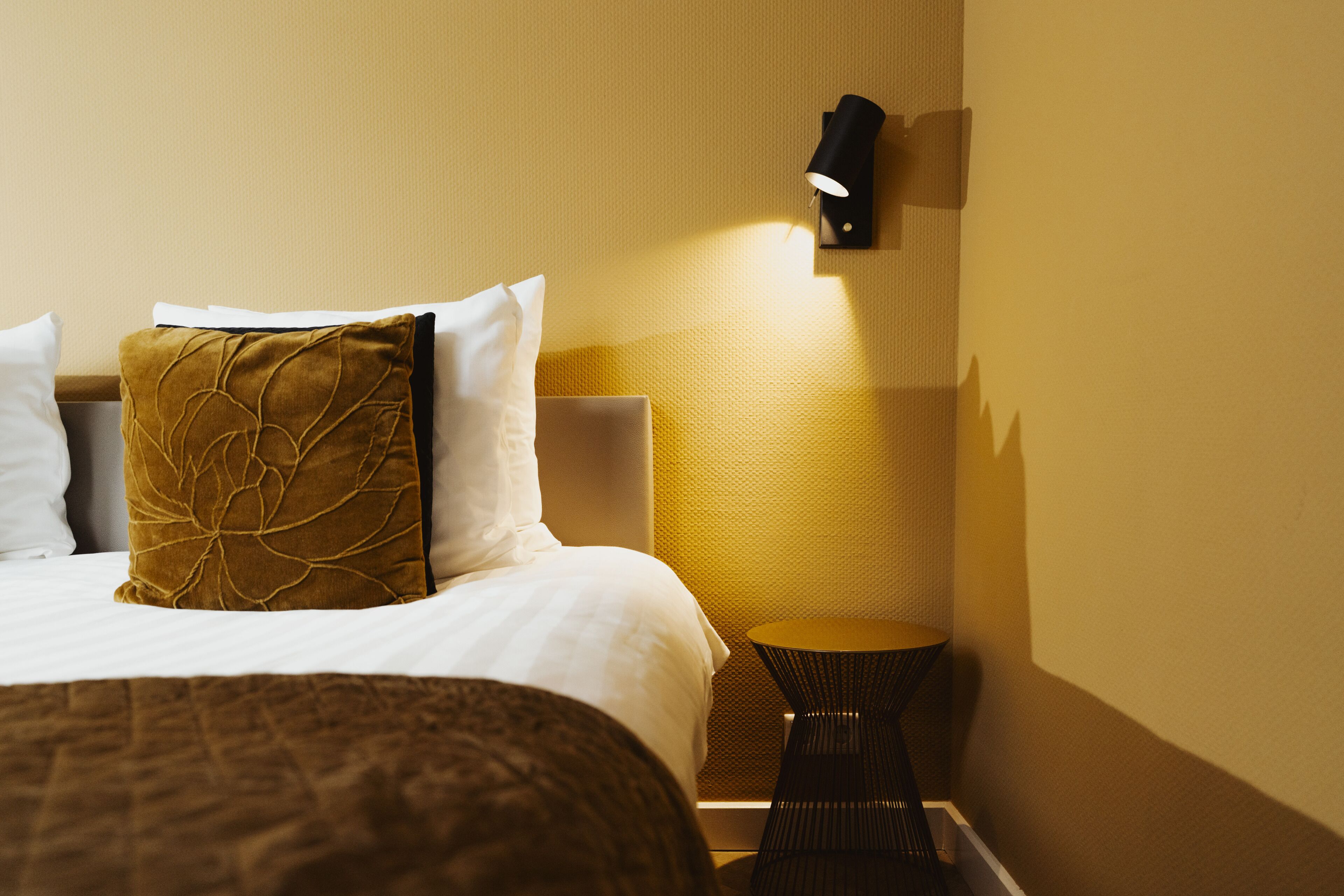 A hotel room with a bed, a nightstand, a lamp, and a wall-mounted light fixture.