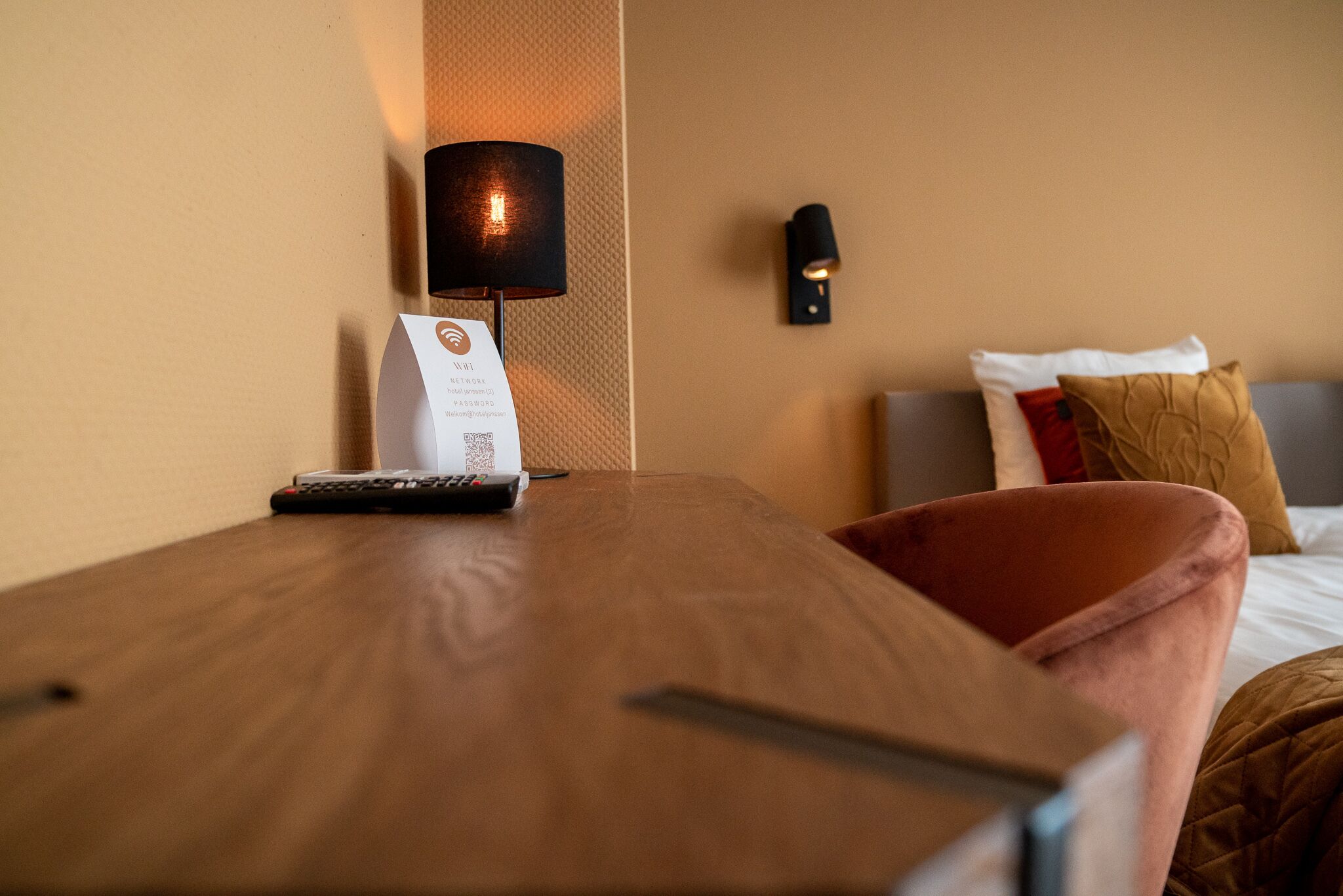 A hotel room with a wooden nightstand, a remote control, a room service card, a lamp, and a bed with pillows.