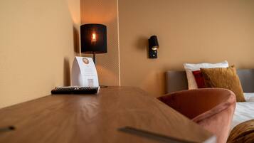A hotel room with a wooden nightstand, a remote control, a room service card, a lamp, and a bed with pillows.