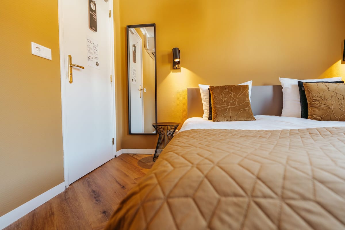 Une chambre d’hôtel avec un lit, une table de chevet, une lampe fixée au mur et une porte dotée d’un miroir.