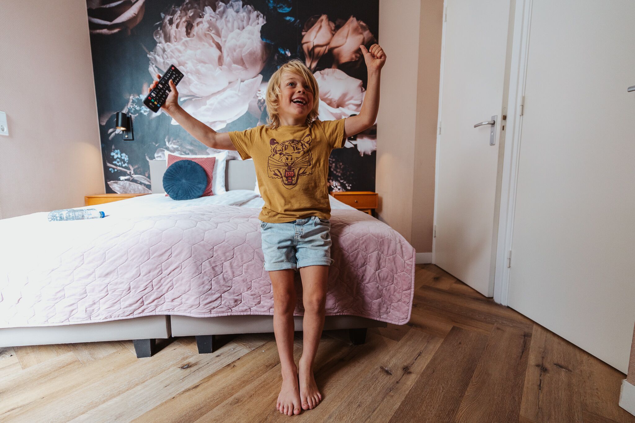 A child sitting on a bed in a room with a large floral wall mural, a pink bedspread, and a wooden floor.