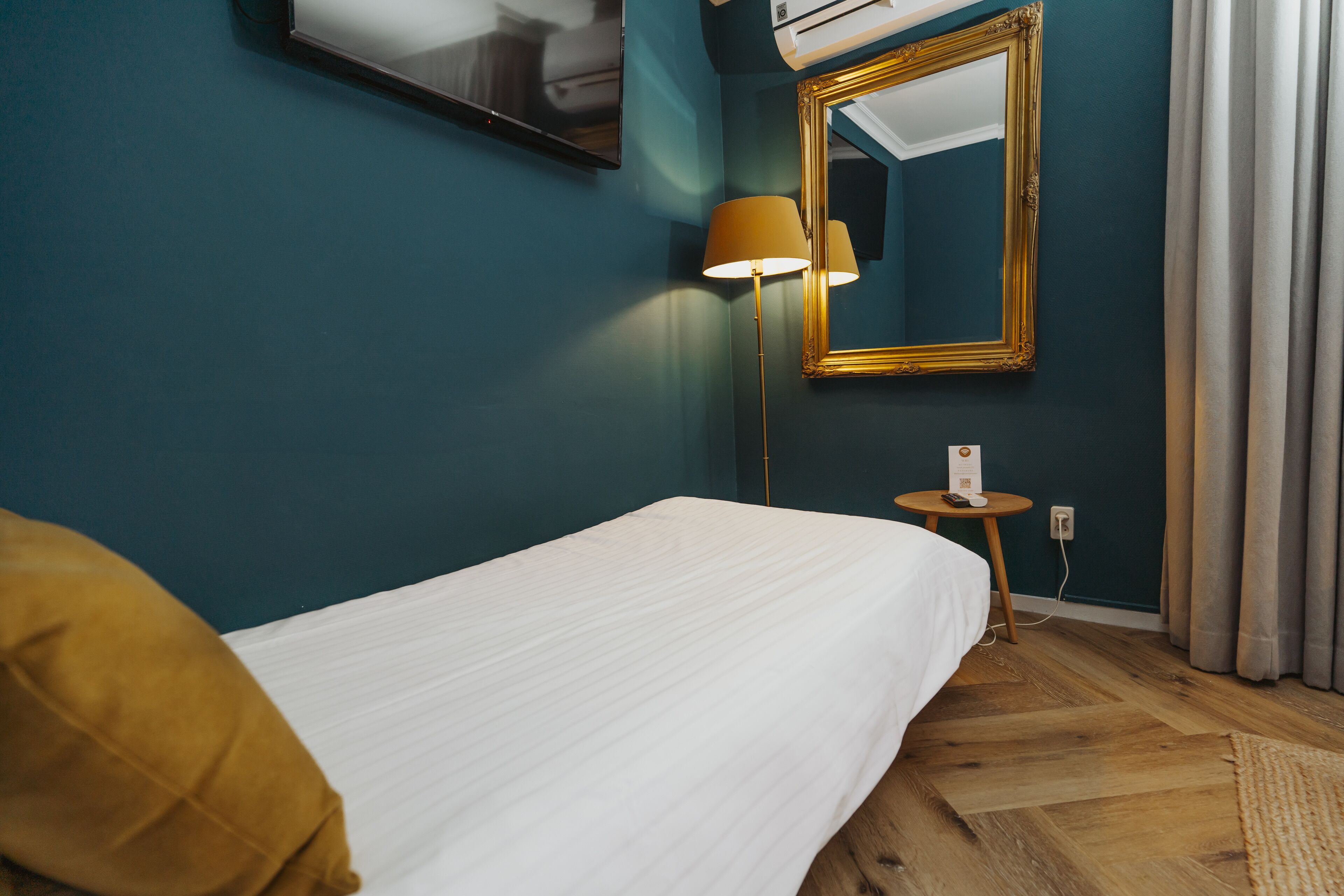 Una habitación de hotel con paredes color turquesa, una cama con ropa blanca, un espejo con marco dorado, una lámpara y una mesita con un teléfono.