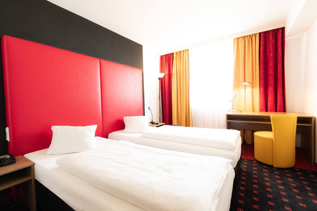 A hotel room with two single beds, a desk with a chair, and a desk lamp.