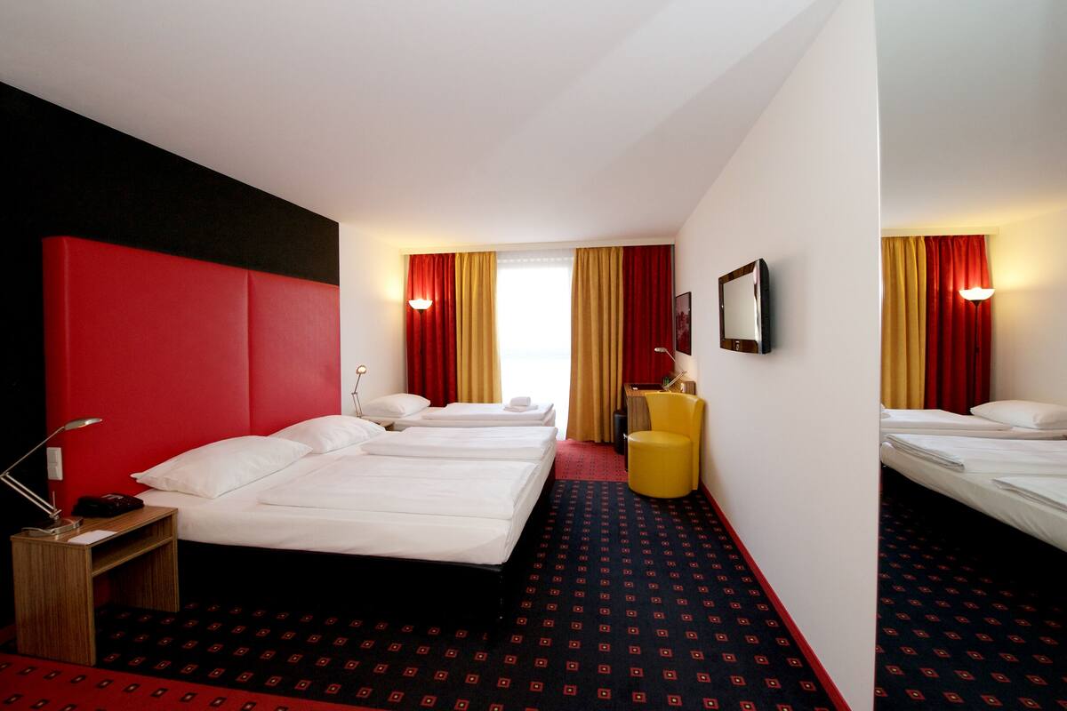 A hotel room with two beds, a red accent wall, a yellow chair, and a desk with a lamp.