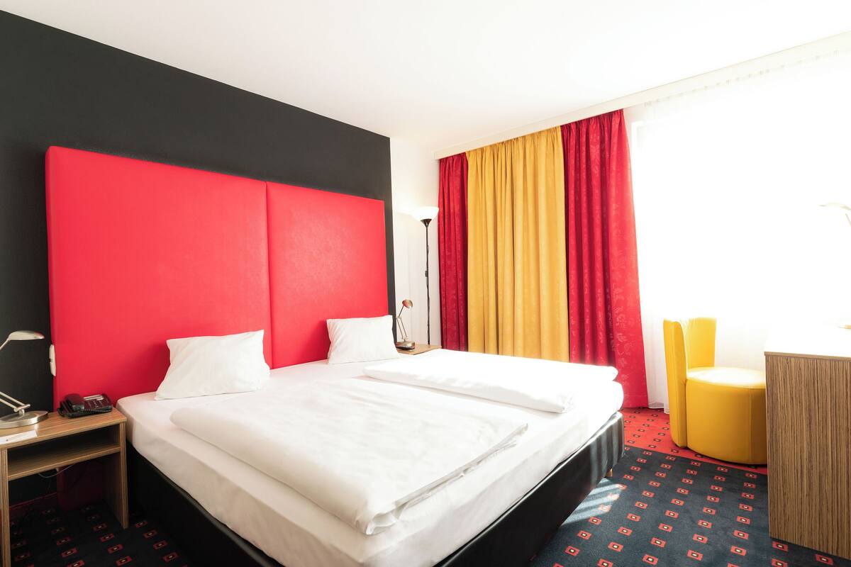A hotel room with a large bed, a desk, a chair, and a window with curtains.