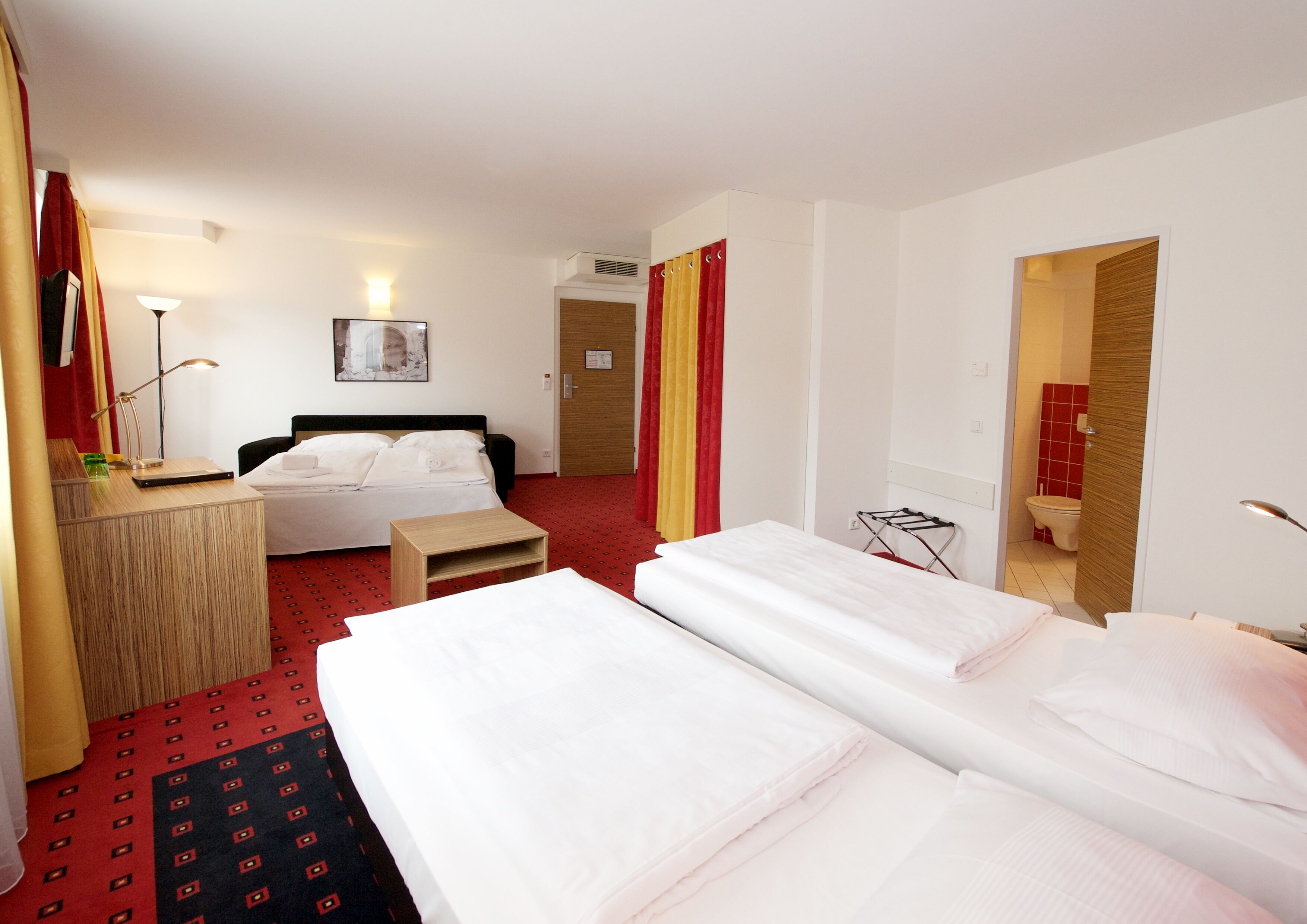 Una camera d'albergo con un letto, una scrivania, una sedia, una lampada e un bagno visibile attraverso una porta aperta.