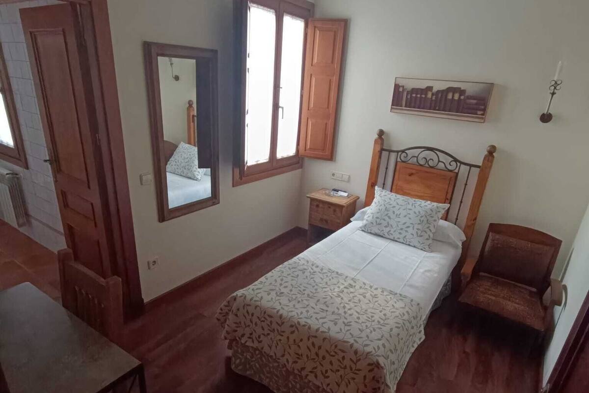 A bedroom with a bed, a nightstand, a chair, a wooden table, a mirror, and a window.
