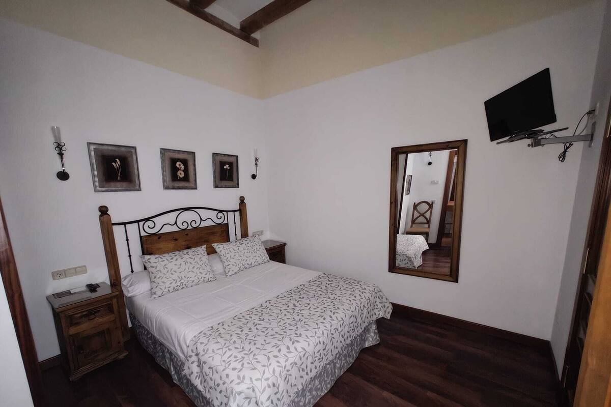 A bedroom with a wooden bed, a nightstand, a bedside table, a mirror, and a TV mounted on the wall.