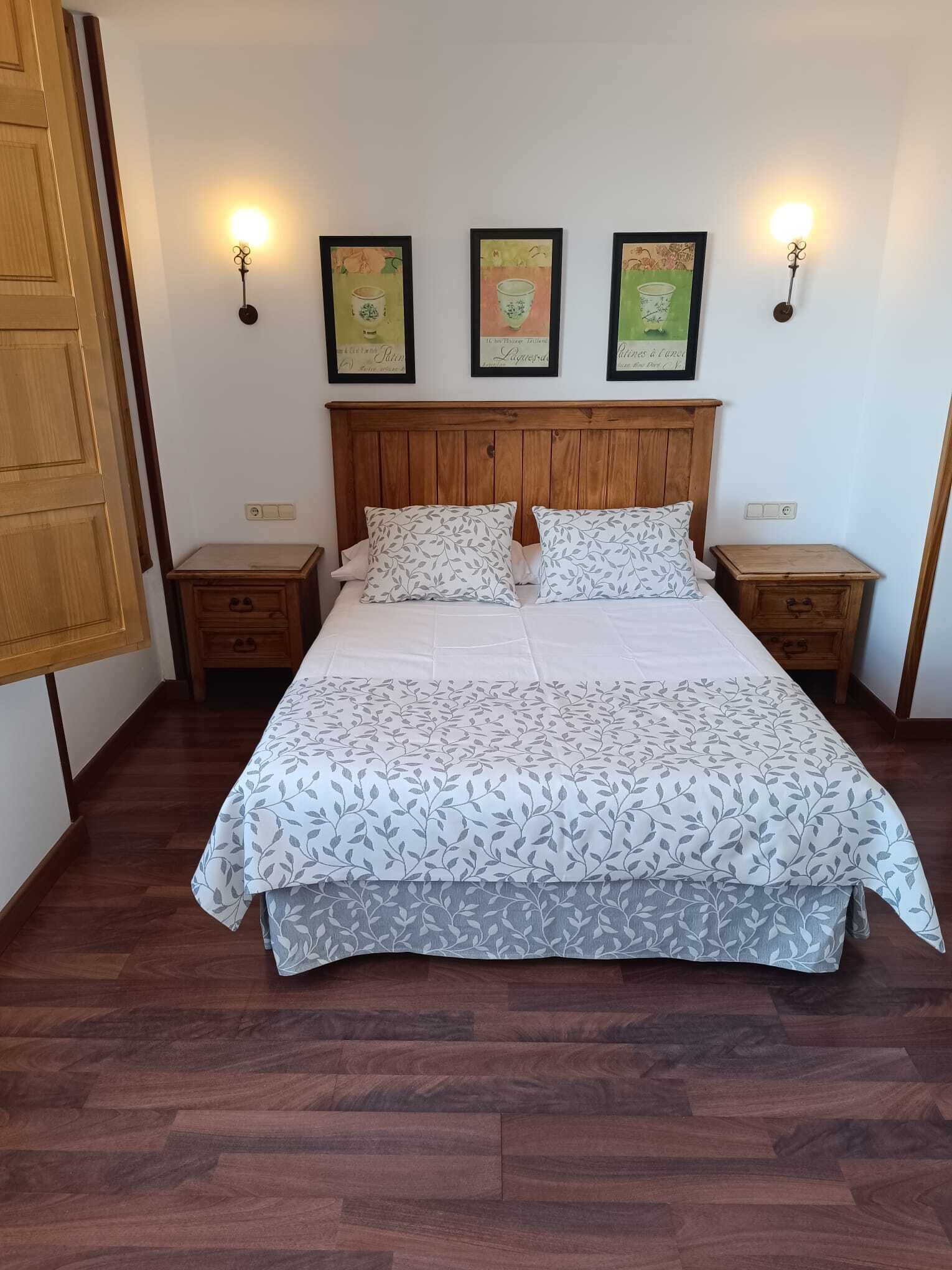 A bedroom with a wooden headboard, a bed with patterned bedding, two bedside tables, and three wall-mounted lamps.