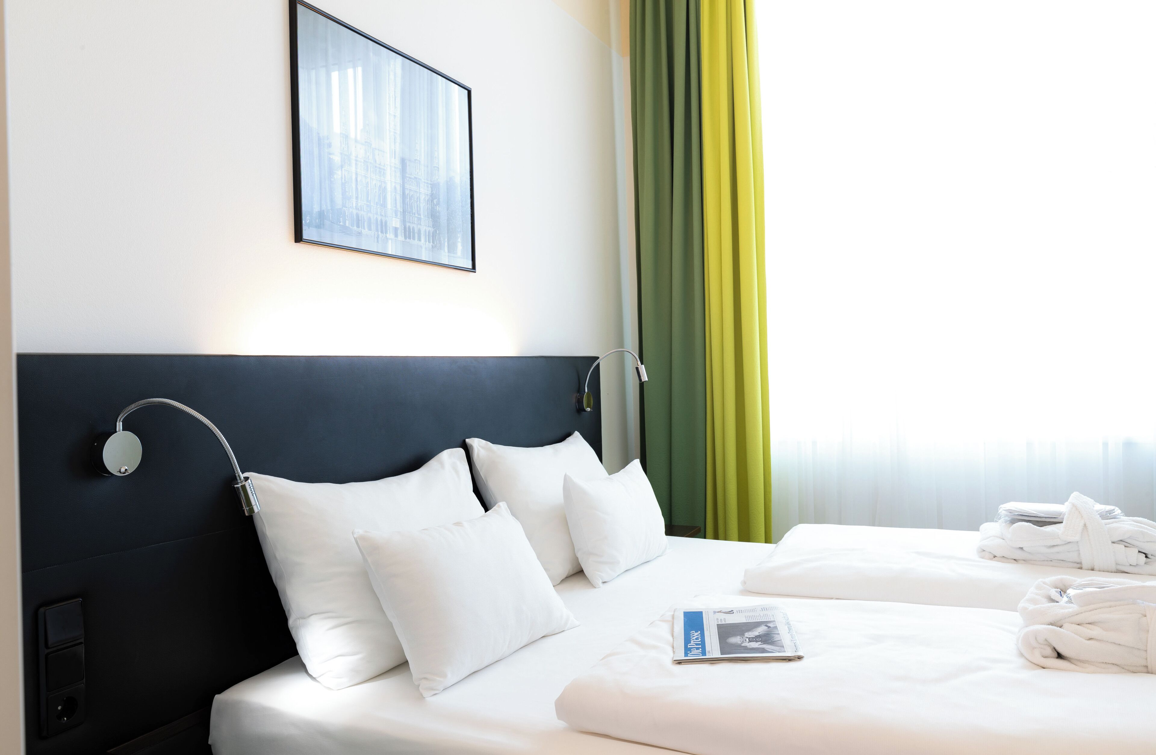 A hotel room with a bed, pillows, a headboard, a framed picture, a window with curtains, and a magazine on the bed.