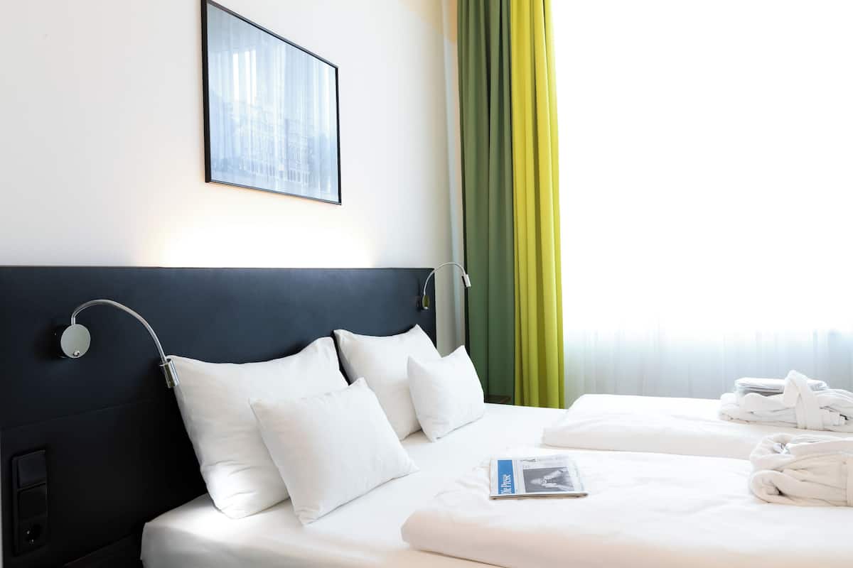 Ein Hotelzimmer mit Bett, Kissen, Kopfteil, einem gerahmten Bild, einem Fenster mit Vorhängen und einer Zeitschrift auf dem Bett.