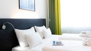 Ein Hotelzimmer mit dunkelbrauner Kopfteil, weißer Bettwäsche, einem gerahmten Bild an der Wand und einem Fenster mit grünen Vorhängen.