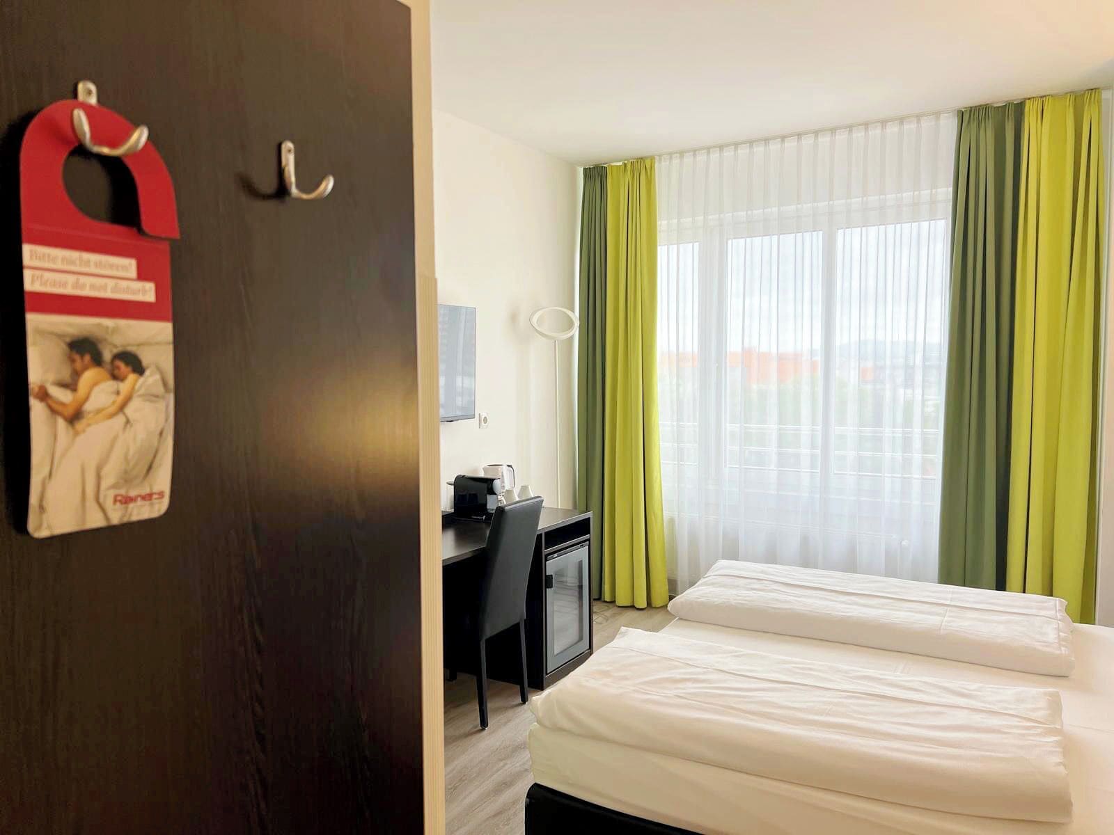 A hotel room with a bed, a desk, and a chair. There is a window with green curtains.