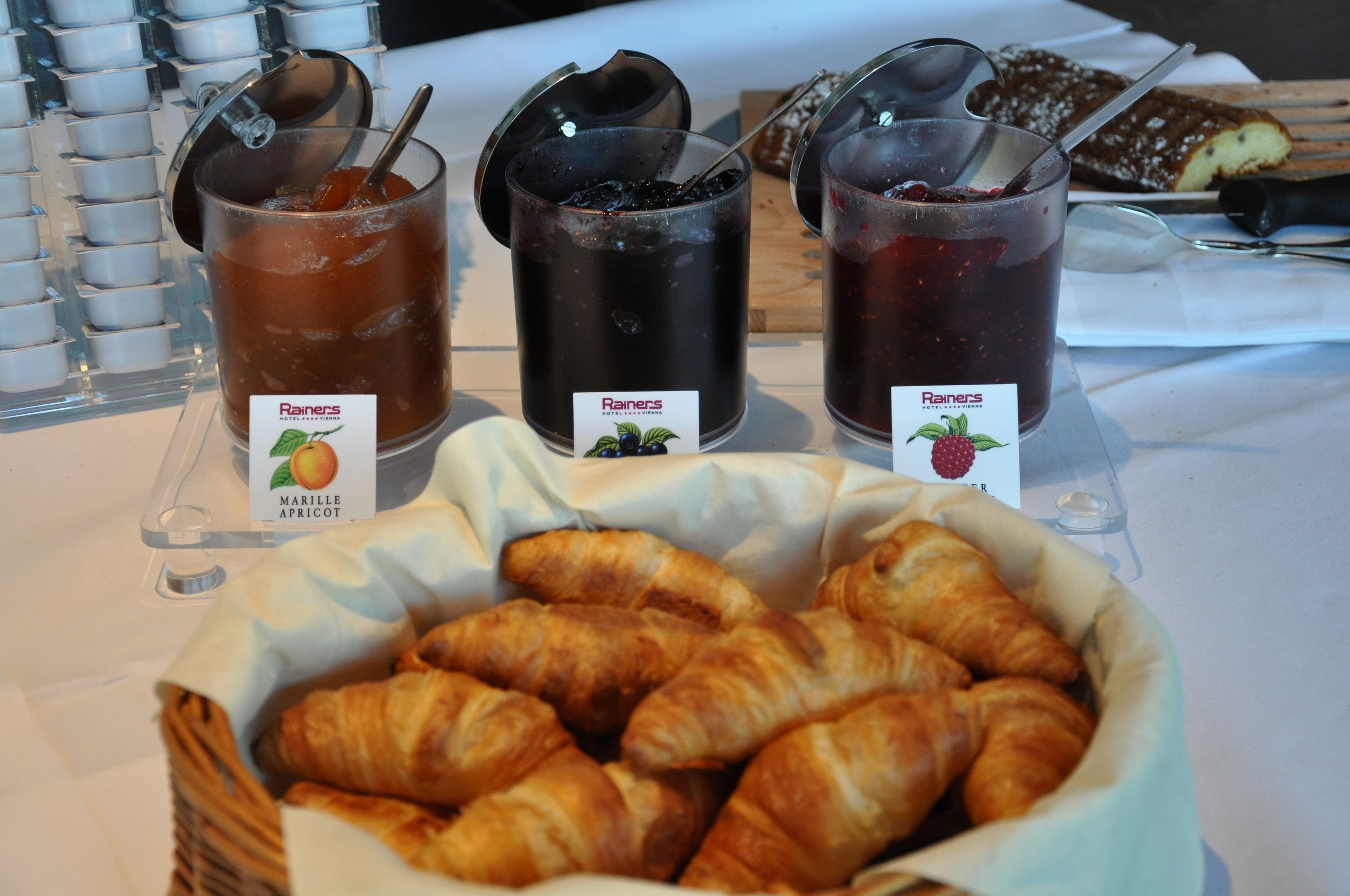 A table with three containers of Rainer's fruit preserves and a basket of croissants.