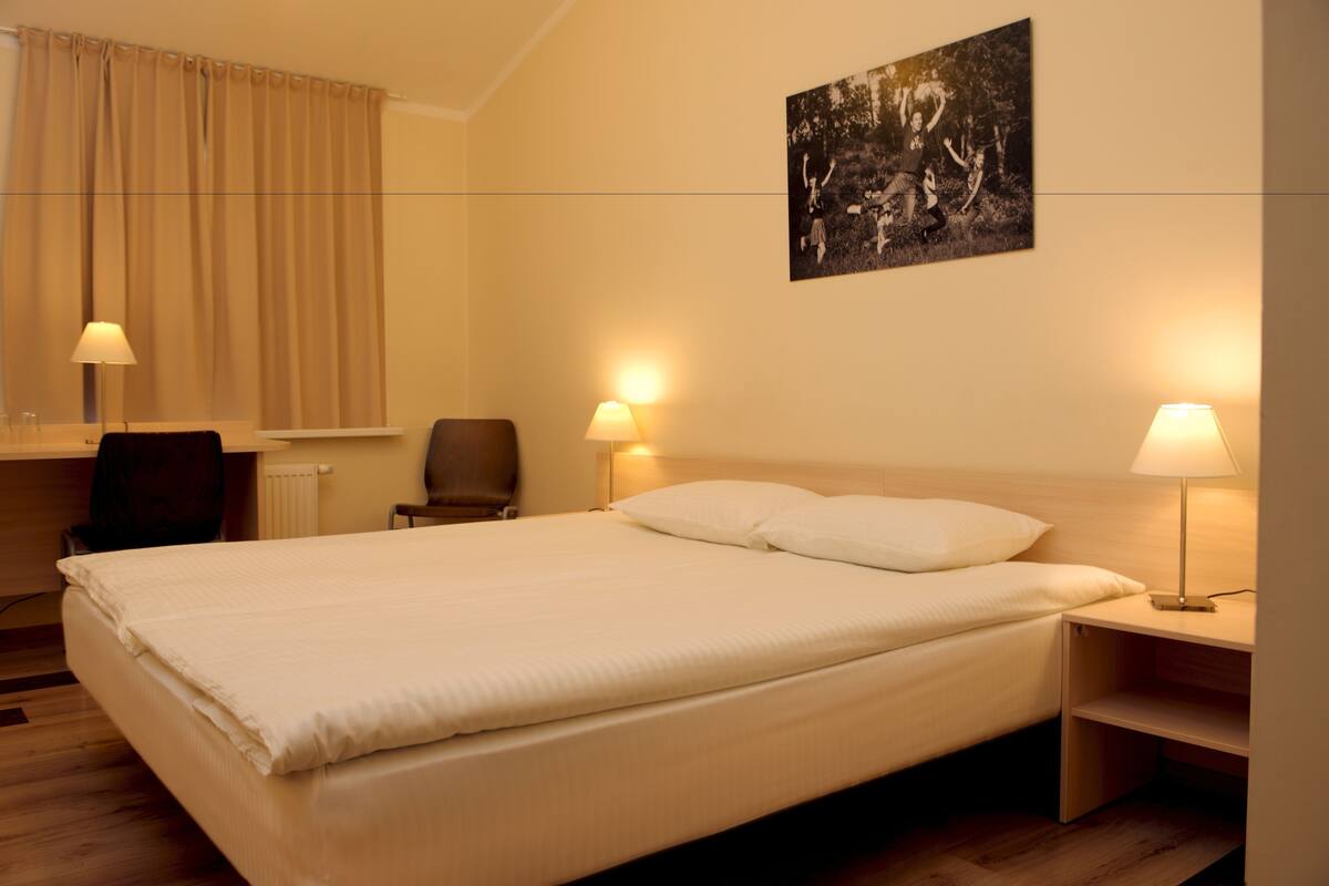Ein Hotelzimmer mit einem großen Bett, zwei Nachttischen mit Lampen, einem Sessel, einem Schreibtisch und einem Fenster mit Vorhängen.