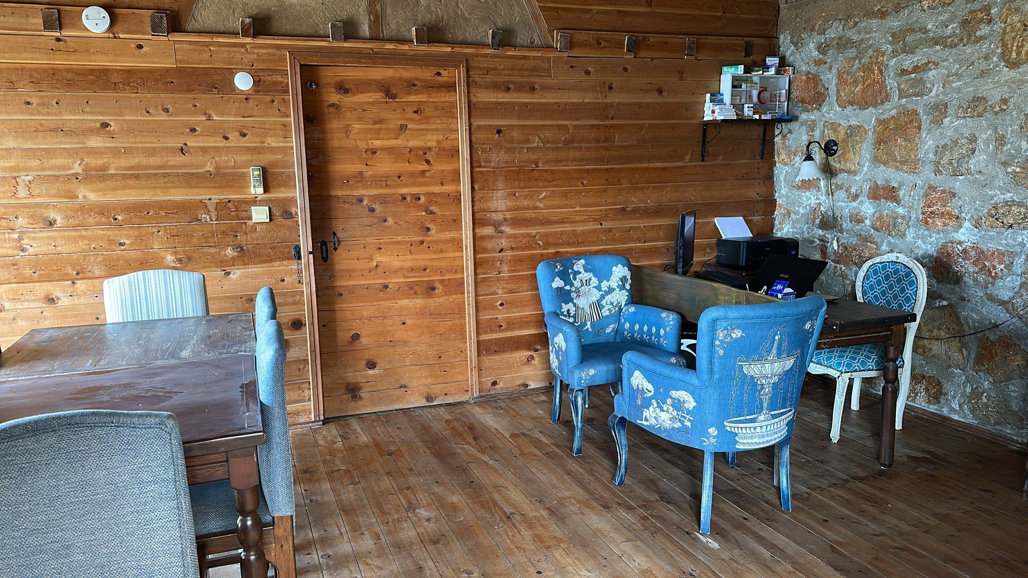 Una habitación rústica de madera con una mesa de comedor, dos sillas tapizadas en azul, una computadora portátil sobre un escritorio de madera y una pared de piedra.