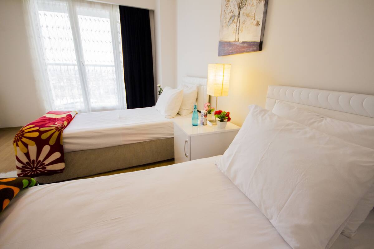 A hotel room with two beds, a nightstand with a lamp, a window with curtains, and a painting on the wall.