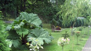 A garden with a wooden cart, large leafy plants, and a weeping willow tree. - Haus Am Wald (Heringsdorf)