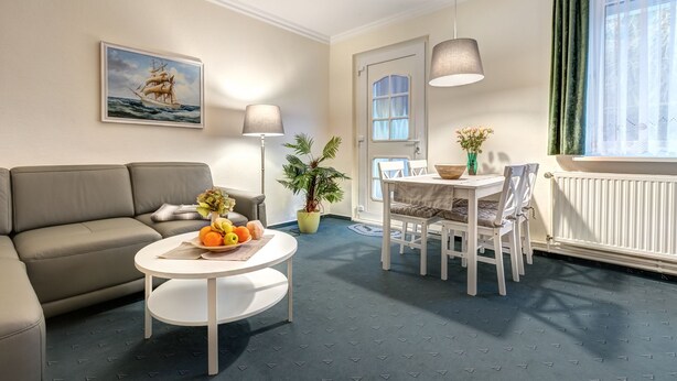 A living room with a sofa, a round coffee table with fruit, a dining table with chairs, a potted plant, and a painting of a ship on the wall. - Haus Am Wald (Heringsdorf)