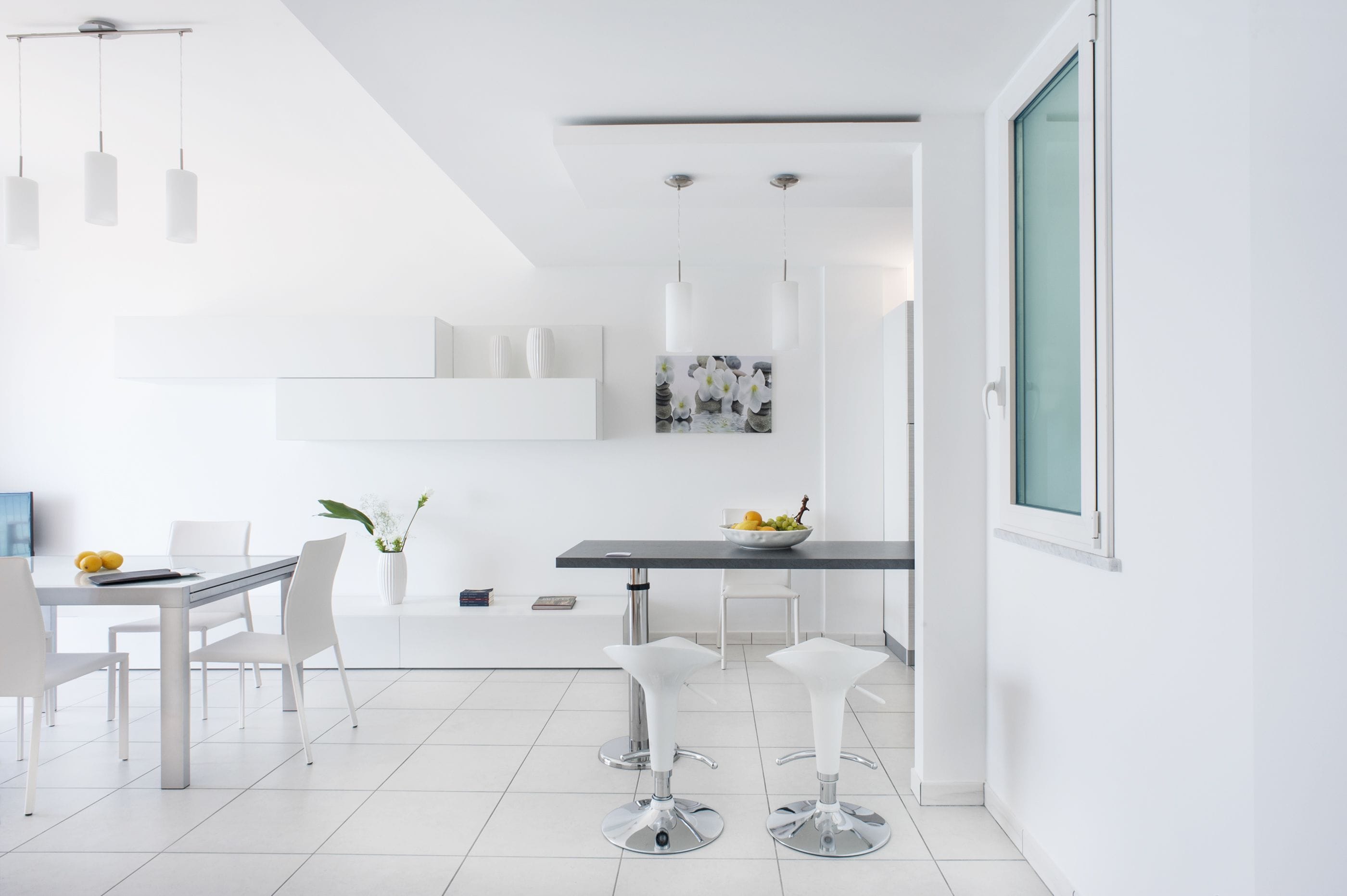 Un espace repas moderne et minimaliste, avec une table blanche, quatre chaises blanches et deux tabourets de bar. On peut voir un saladier de fruits sur la table et un vase avec des fleurs sur le sol.
