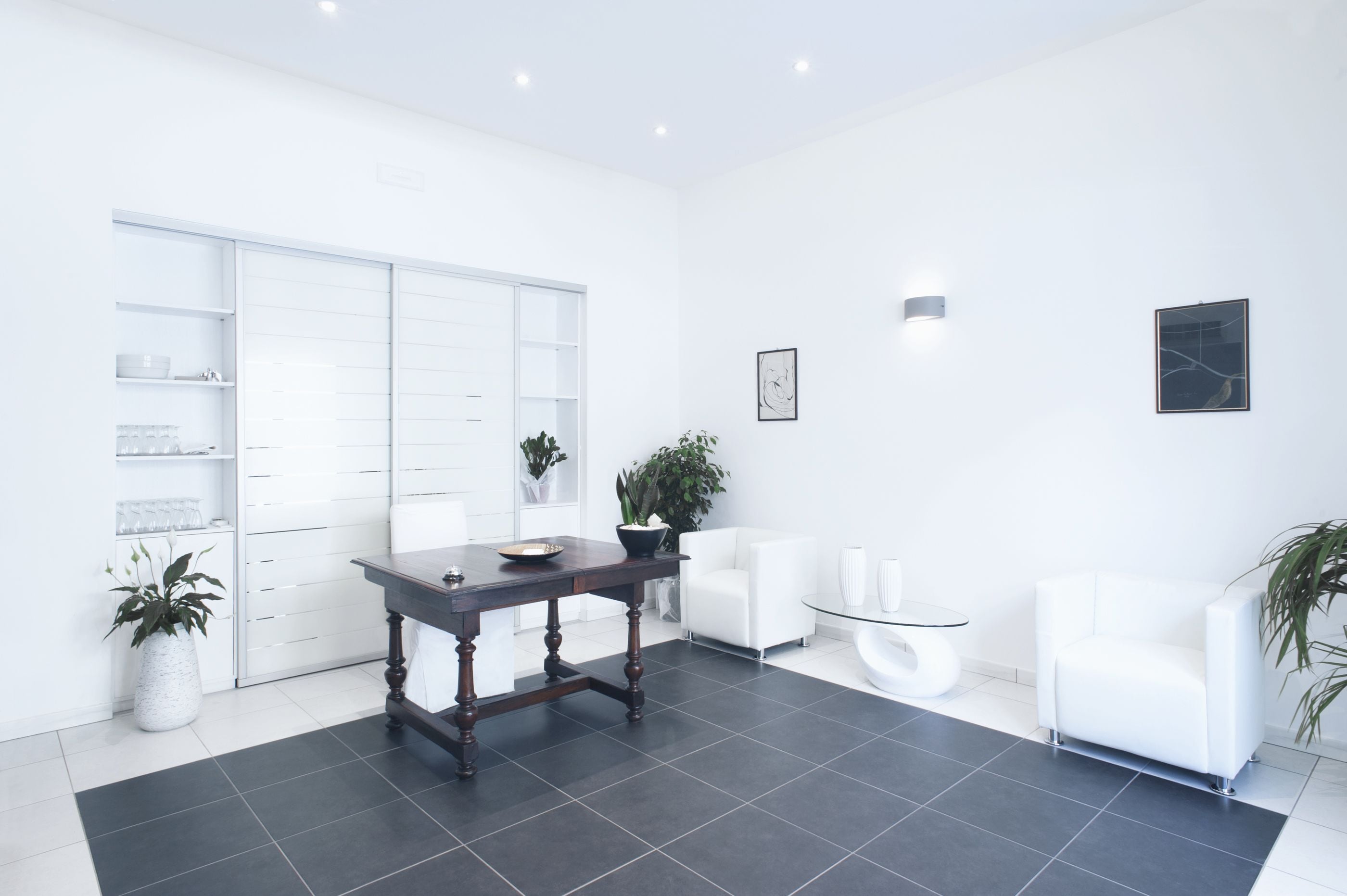 A modern, well-lit room with a dark wooden table, white chairs, a glass coffee table, and a white wardrobe with shelves.