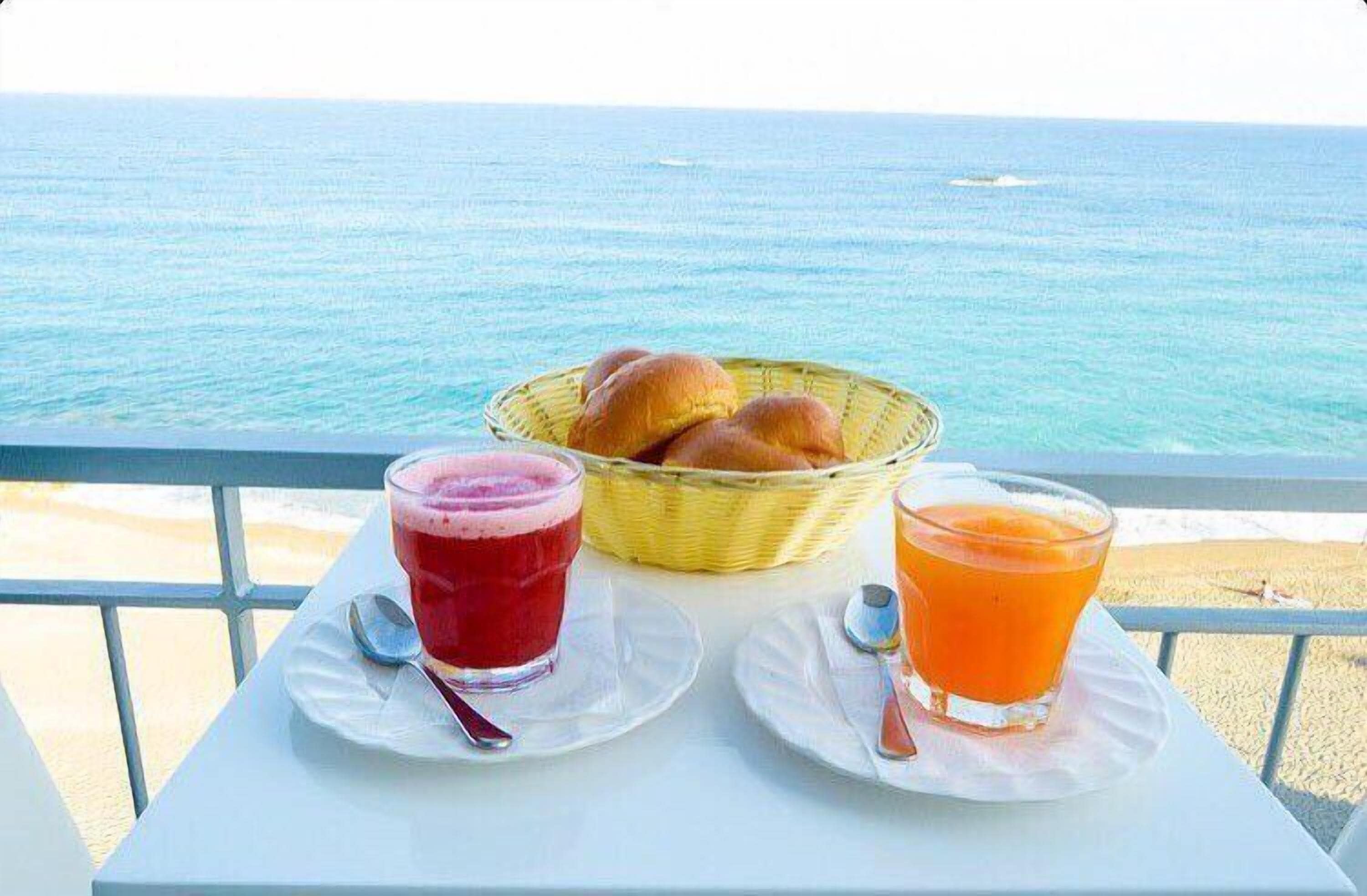 A table with a basket of bread and two glasses of juice, overlooking the sea.
