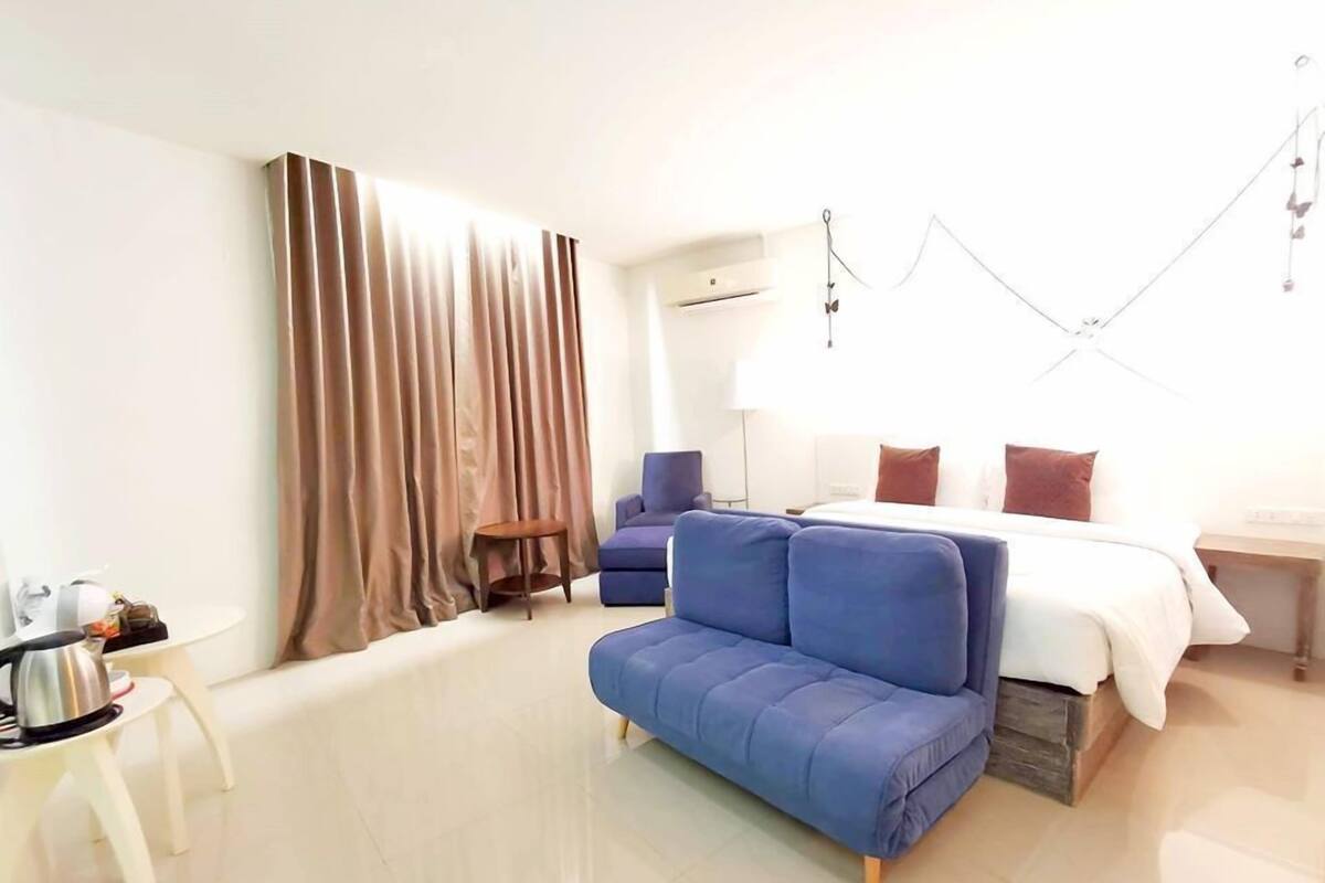 Una habitación de hotel moderna con cama, sofá azul, mesita con tetera y ventana con cortinas.