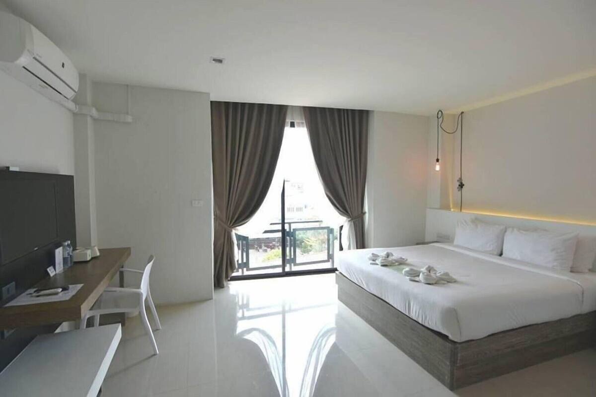 Habitación de hotel con cama, escritorio, silla, televisor y balcón con cortinas.