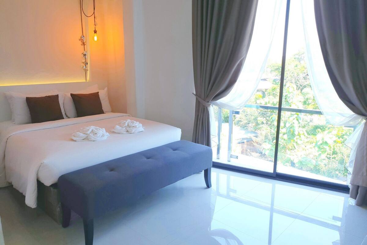 Habitación de hotel moderna con una cama grande, un banco y vistas al exterior con vegetación.