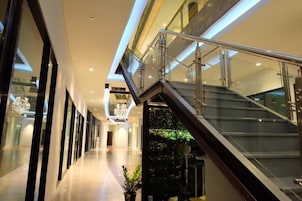 Un corredor interior moderno con barandilla de cristal, una escalera, una araña y una planta en maceta.