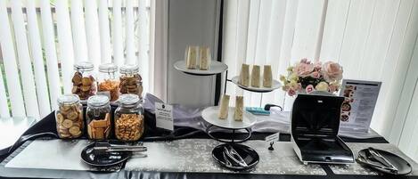Una mesa de buffet con diversos alimentos, incluyendo frascos de nueces y galletas, un soporte para pasteles con cupcakes y una máquina de café.
