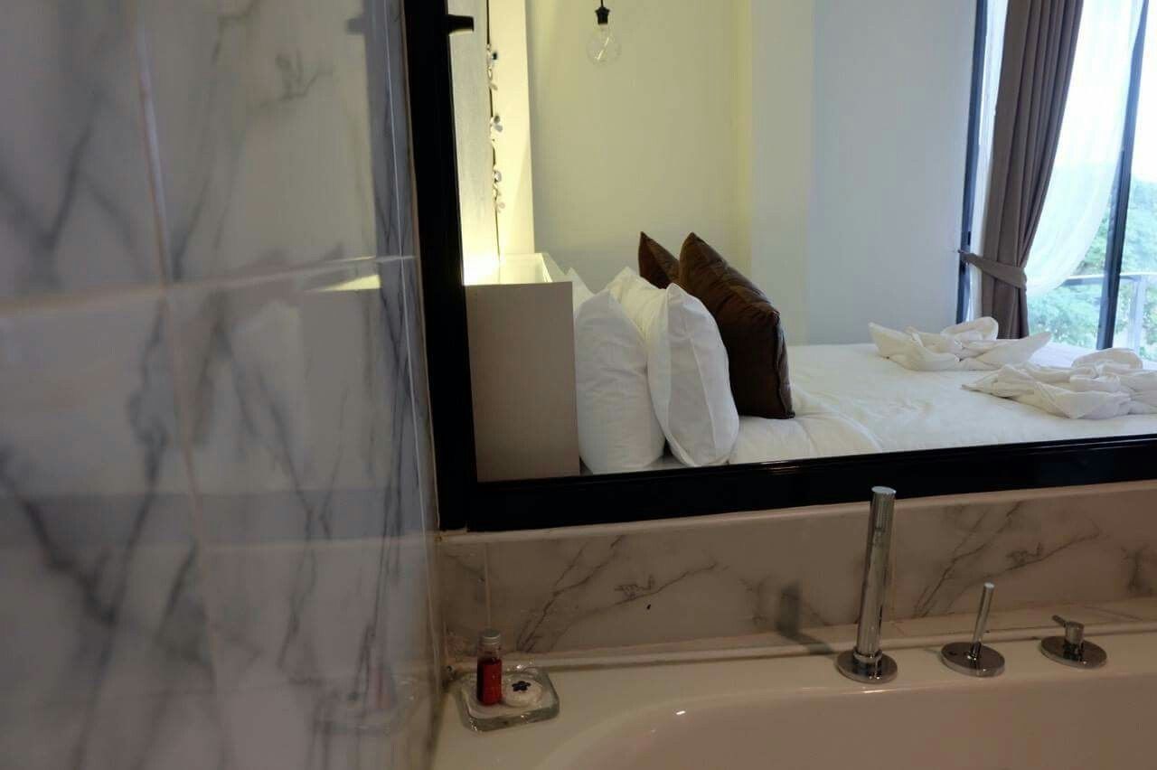 Une chambre d’hôtel avec un grand miroir, un lit recouvert de draps blancs, un mur recouvert de carreaux de marbre et une fenêtre avec des rideaux.