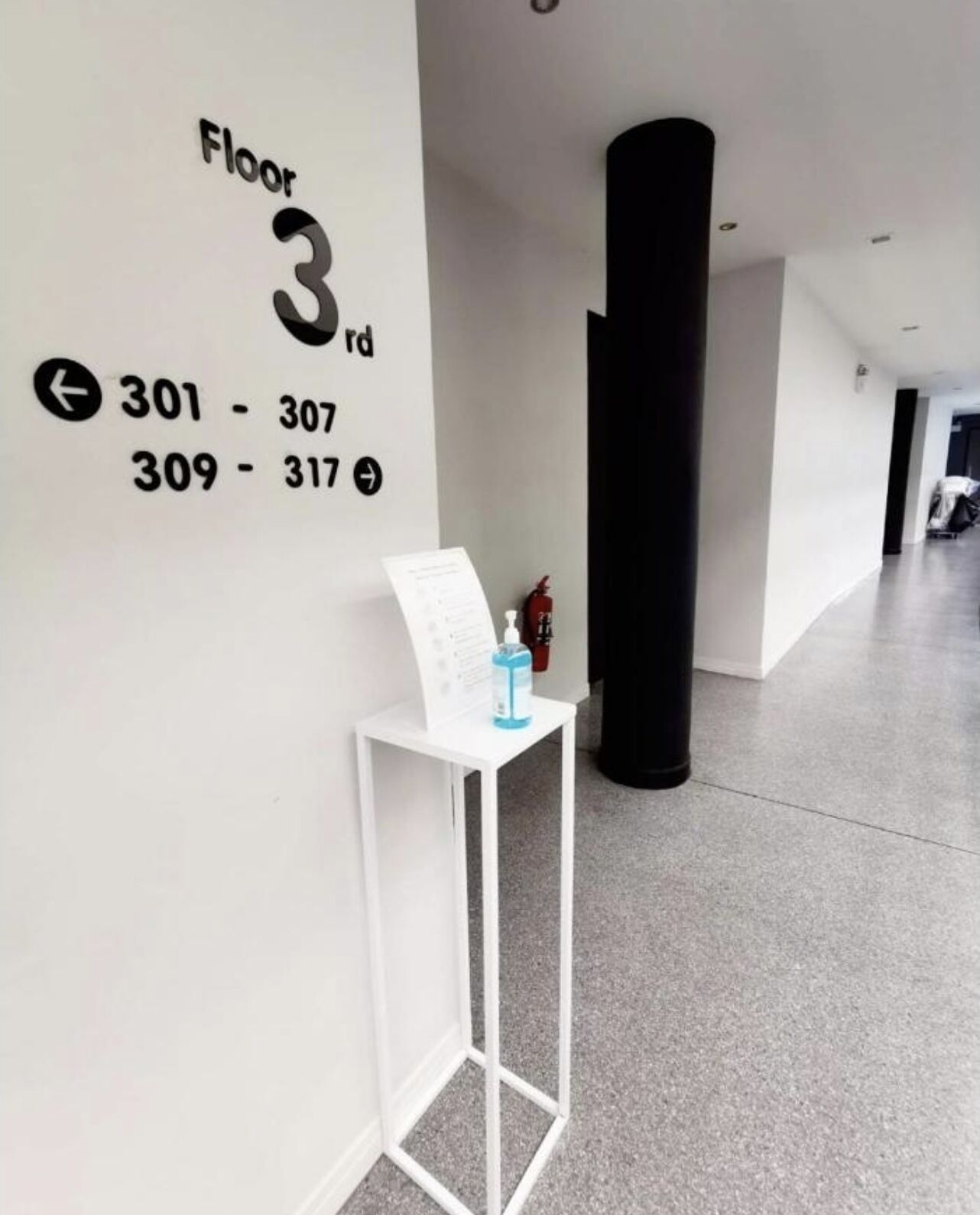 Un pasillo con un cartel que indica 'Floor 3rd' y números de habitación '301-307' y '309-317'. Se ve una mesa blanca con un objeto azul encima.