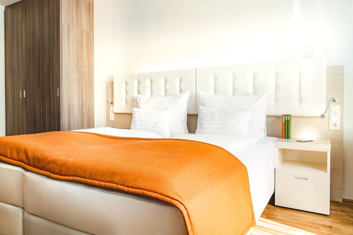 Uma cama bem arrumada com roupa de cama branca e um cobertor laranja, uma cabeceira branca, uma mesa de cabeceira com abajur e um guarda-roupa de madeira.