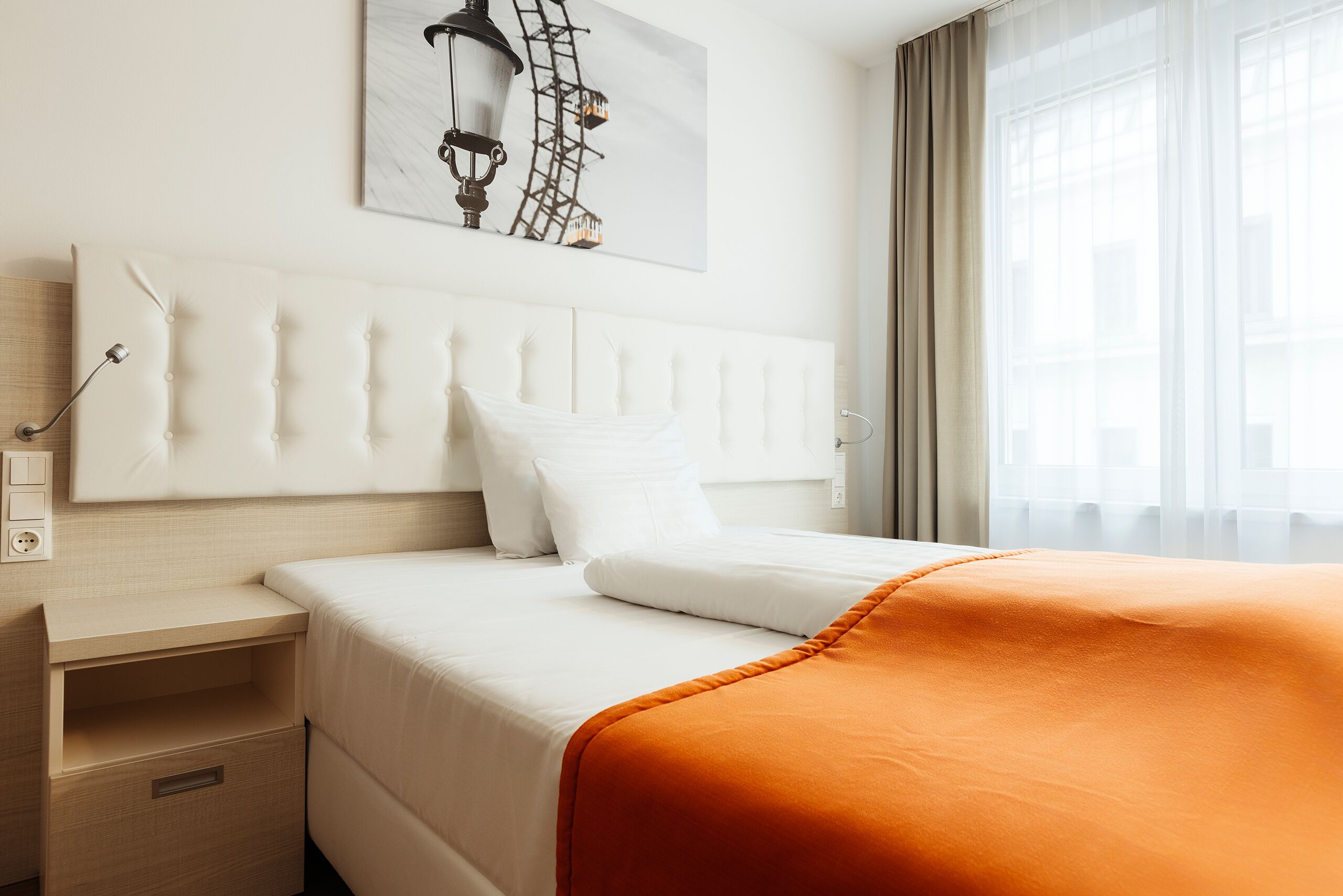 Een hotelkamer met een bed, nachtkastje, een lampje aan de muur en een raam met gordijnen.