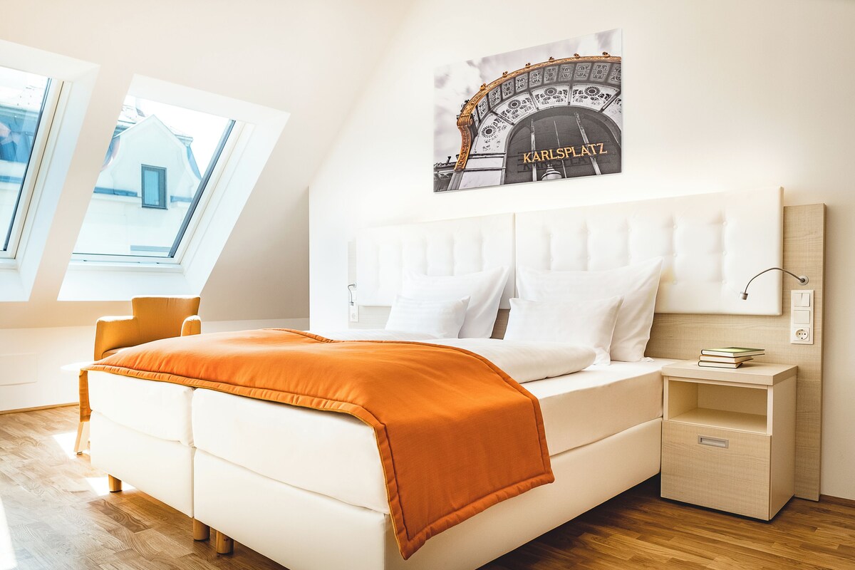 A modern bedroom with a white bed, an orange throw blanket, a headboard with a light switch, and a framed picture of a building on the wall.