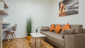 A modern living room with a grey sofa, orange cushions, a white coffee table, a tall green plant, a grey chair, and a painting of a classical statue.