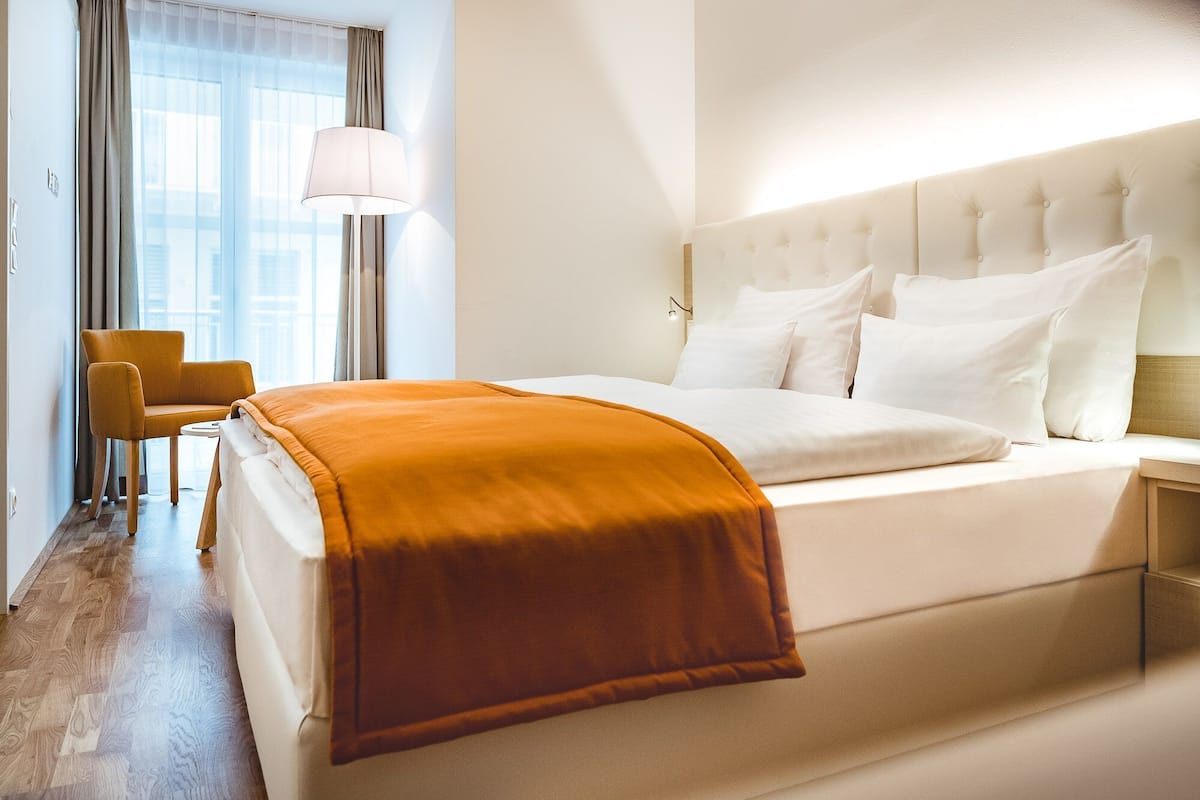 Quarto de hotel com uma cama grande, uma luminária, uma cadeira e vista para o exterior pela janela.