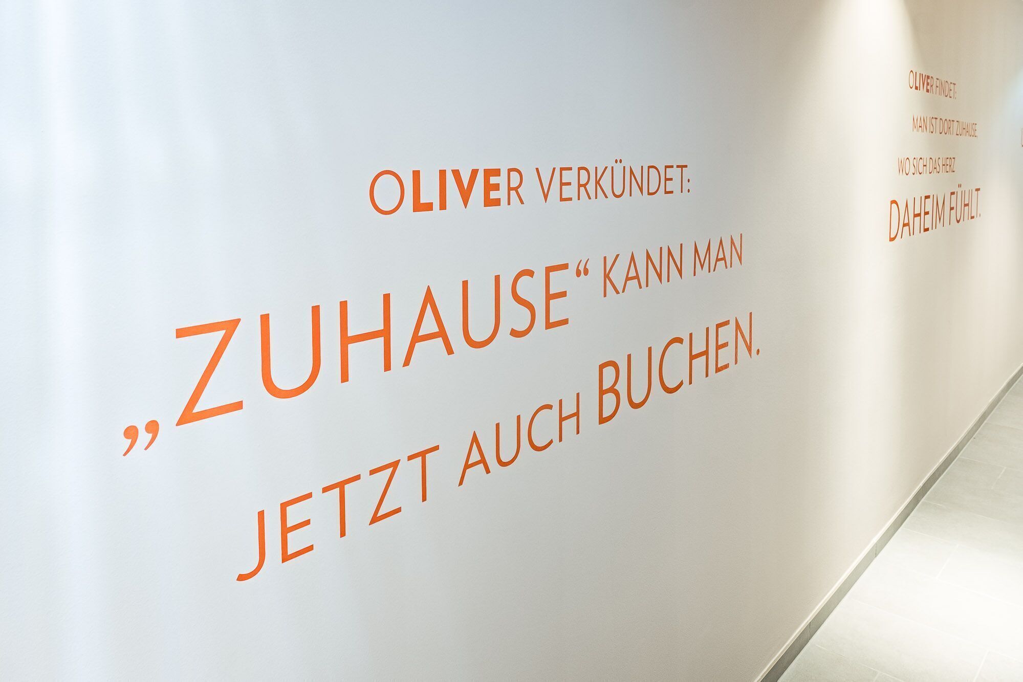 Una pared con texto en alemán, que incluye la frase “OLIVER VERKÜNDET: ZUHAUSE” y “KANN MAN JETZT AUCH BUCHEN”.