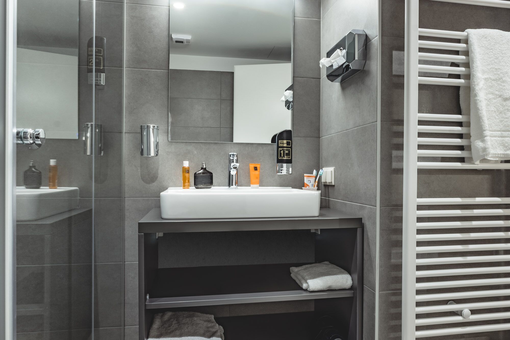 Une salle de bain moderne avec un lavabo blanc, des carreaux gris, un miroir, un porte-serviettes et une étagère sur laquelle est posée une serviette.
