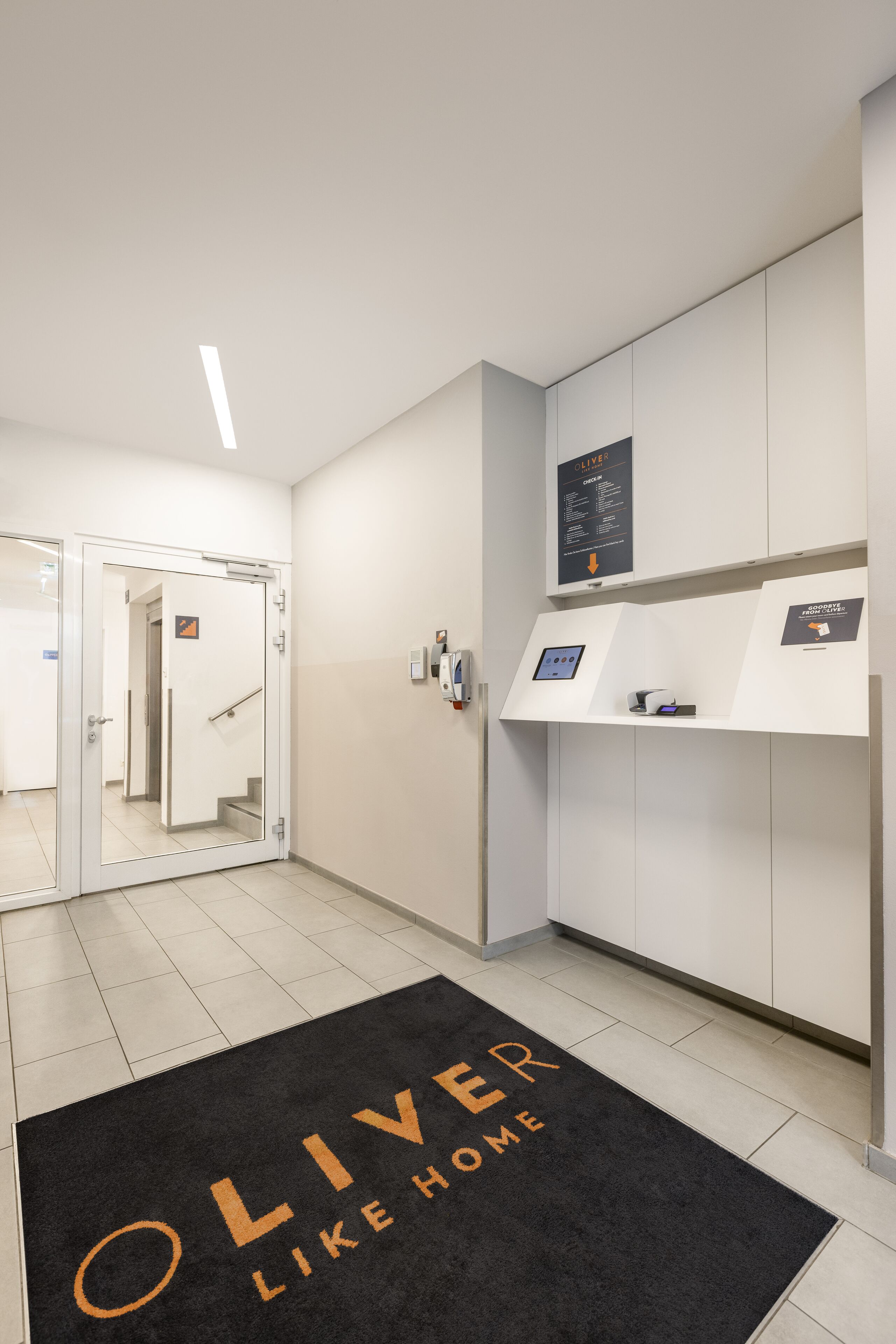 Zona de recepción de hotel con un felpudo negro que dice "OLIVER LIKE HOME", un mostrador de recepción, un quiosco digital y un letrero en la pared.